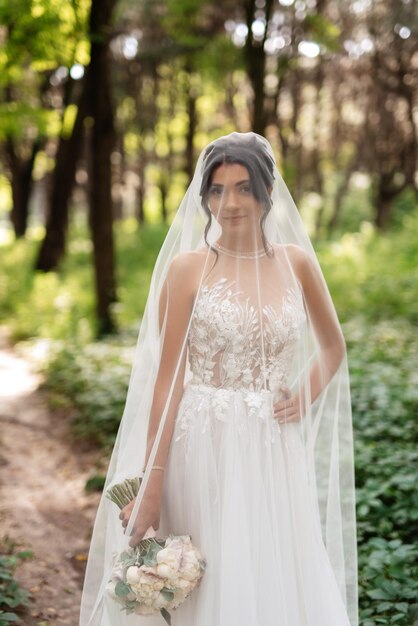 Portrait d'une jeune mariée élégante sur un chemin dans une forêt de feuillus avec un bouquet