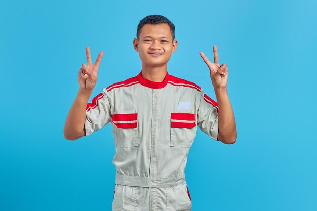 Portrait de jeune mécanicien asiatique souriant faisant signe de paix avec le doigt isolé sur fond bleu