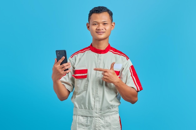 Portrait de jeune mécanicien souriant, pointant le doigt sur un téléphone portable isolé sur fond bleu