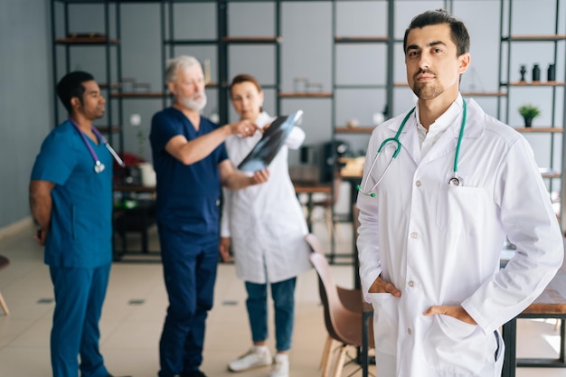 Portrait d'un jeune médecin confiant en uniforme blanc debout dans un bureau de réunion médicale, regardant la caméra. Équipe multiethnique de médecins travaillant en arrière-plan, discutant de l'IRM du patient.
