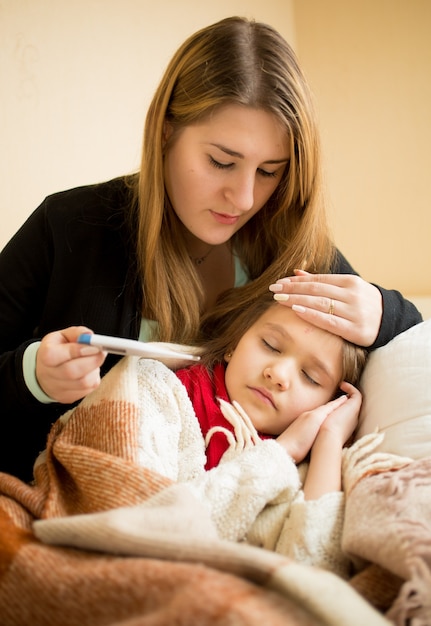 Portrait de jeune mère embrassant sa fille malade et vérifiant le thermomètre