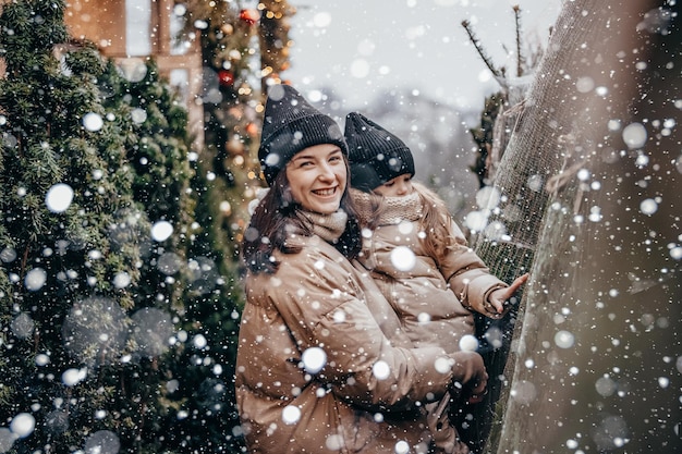 Portrait d'une jeune mère avec sa fille dans ses bras, ils sourient et se réjouissent des chutes de neige Choisir et acheter un sapin de Noël au marché de Noël