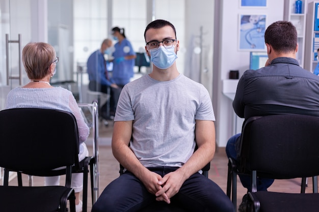 Portrait d'un jeune patient souriant regardant la caméra assis sur une chaise dans la salle d'attente de la clinique de l'hôpital en respectant la distanciation sociale pendant que le médecin travaille en arrière-plan