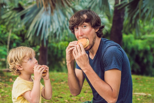 Portrait d'un jeune père et de son fils dégustant un hamburger dans un parc et souriant.