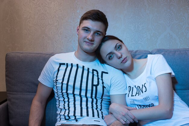 Portrait de jeune et romantique couple assis en embrassade sur un canapé ensemble devant une télévision à la maison dans une atmosphère détendue et regardant la télévision