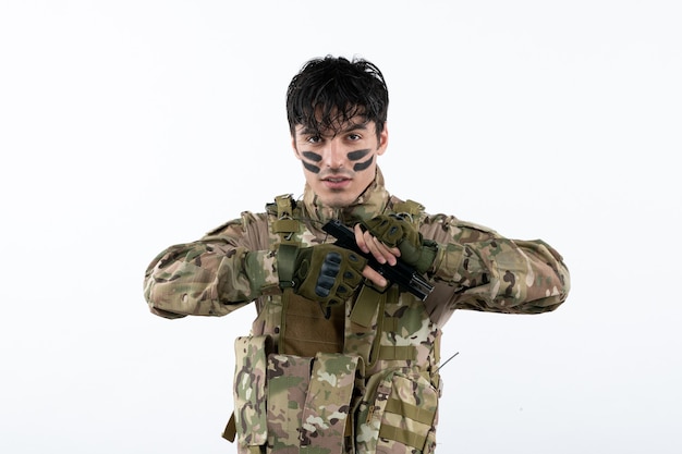 Portrait de jeune soldat en tenue de camouflage avec pistolet sur mur blanc