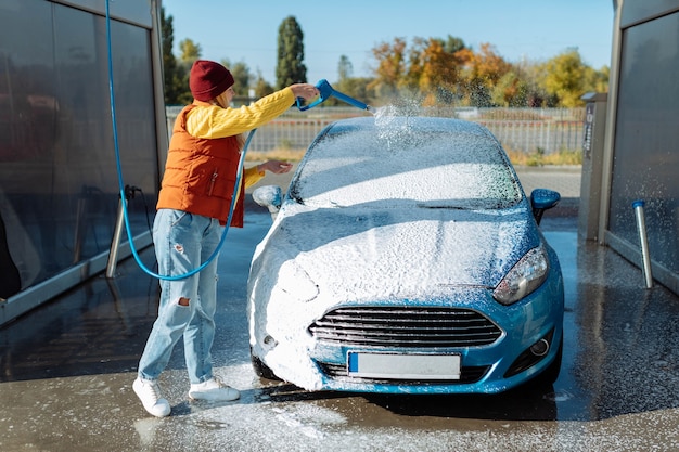 Portrait jeune, souriante, heureuse, jolie femme lavant l'automobile à la station de lavage de voiture manuelle en libre-service, nettoyant avec de la mousse, de l'eau sous pression. Transport, automobile, concept d'entretien des véhicules.