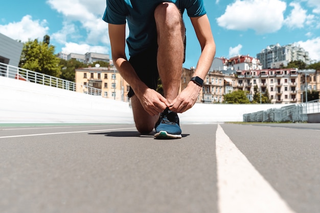 Portrait de jeune sportif athlétique en smartwatch touchant des baskets sur une piste de course avec des bâtiments