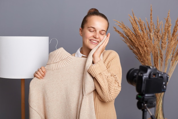 Portrait d'une jeune styliste blogueuse satisfaite et ravie avec une coiffure en chignon filmant une vidéo sur un trépied montrant aux abonnés un pull chaud et doux avec une nouvelle collection