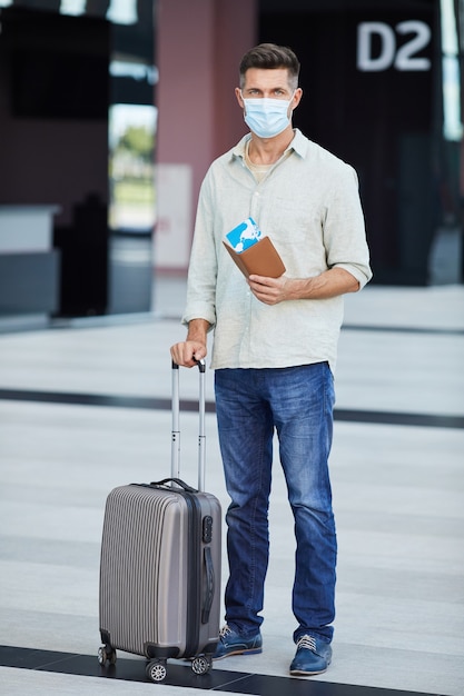 Portrait de jeune touriste en masque de protection avec des bagages et des billets à la recherche en se tenant debout à l'aéroport pendant la pandémie