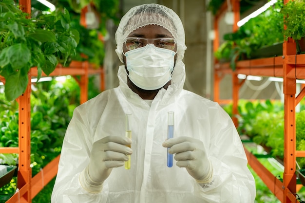 Portrait de jeune travailleur de culture afro-américain en casquette, masque et lunettes de sécurité debout avec des tubes à essai de liquides colorés à la ferme verticale