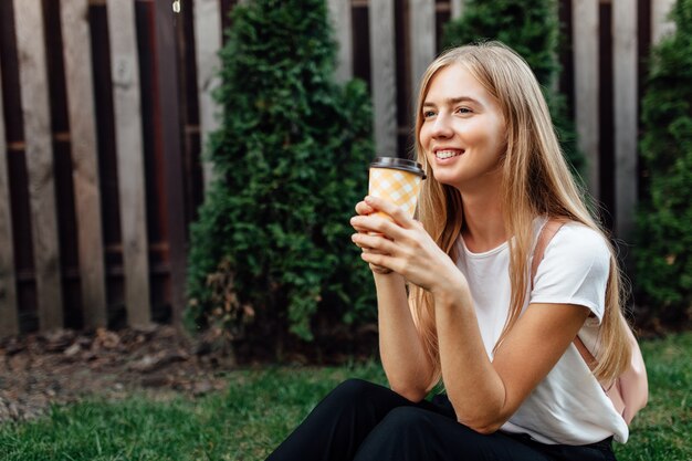 Portrait d'un jeune, vêtu d'un t-shirt blanc, à l'extérieur. Fille assise sur la pelouse et boire du café.