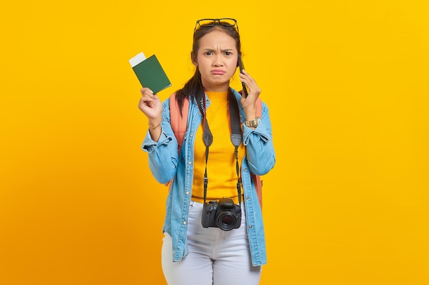 Portrait d'un jeune voyageur asiatique pensif tenant un billet de passeport de carte d'embarquement et parlant sur un smartphone isolé sur fond jaune. Passagers voyageant le week-end. Concept de voyage en avion