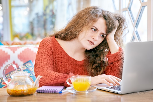 Portrait de jeunes étudiantes blanches avec des taches de rousseur aux longs cheveux roux bouclés travaillant avec un ordinateur portable avec une expression frustrée et fatiguée.