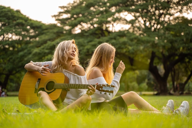 Photo portrait de jeunes femmes de race blanche assis dans le parc en plein air et jouer de la guitare chanter une chanson avec bonheur