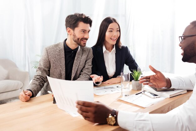 Portrait de jeunes hommes d'affaires et femmes d'affaires multinationaux souriants assis à la table et discutant d'un nouveau projet tout en travaillant ensemble au bureau