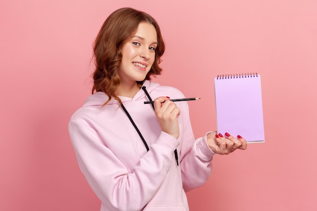 Portrait d'une jolie adolescente aux cheveux bouclés en sweat à capuche pointant un crayon sur une feuille de cahier vide et souriant sincèrement devant la caméra, liste de tâches. Studio intérieur tourné isolé sur fond rose
