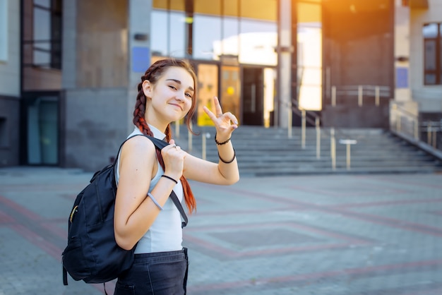 Portrait de jolie adolescente joyeuse en t-shirt blanc avec sac à dos noir sur son épaule. Élève du secondaire élégant fait des gestes avec ses mains sur le fond du bâtiment du Collège.