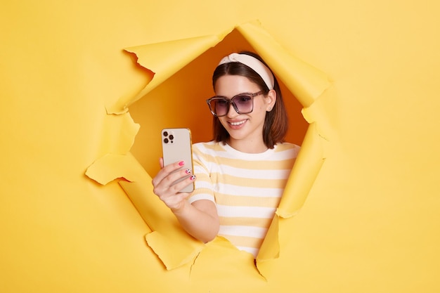 Portrait d'une jolie belle femme à lunettes de soleil se tient dans un trou de papier déchiré à l'aide d'un téléphone portable faisant un selfie ou ayant un appel vidéo debout dans une percée de fond jaune