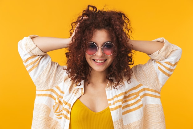 Portrait d'une jolie belle jeune femme bouclée posant isolée sur un mur jaune portant des lunettes de soleil.