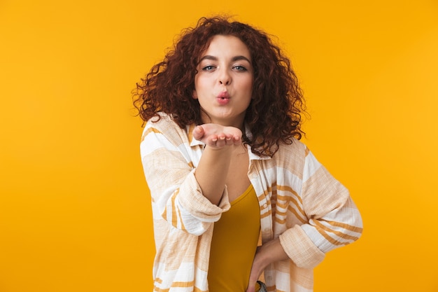 Portrait d'une jolie belle jeune femme bouclée posant isolée sur un mur jaune soufflant des baisers.