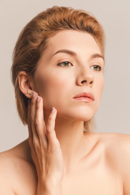 Portrait d'une jolie blonde aux yeux verts et aux cheveux courts. Il touche son visage avec ses mains, un contact doux. Tourné en studio sur fond clair. Maquillage nude. Concept de peau saine