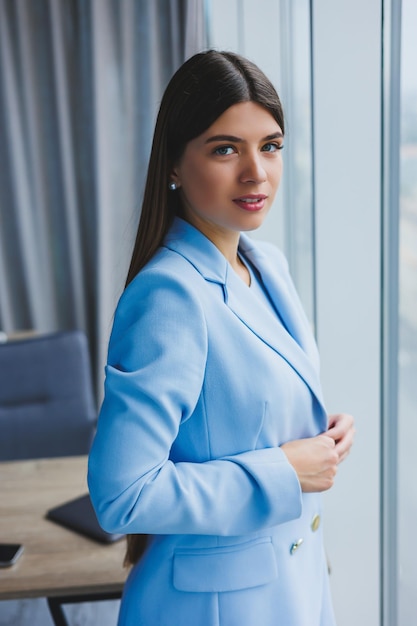 Portrait d'une jolie brune dans une veste bleue au bureau près de la fenêtre panoramique avec vue sur la ville Heureuse femme d'affaires au bureau