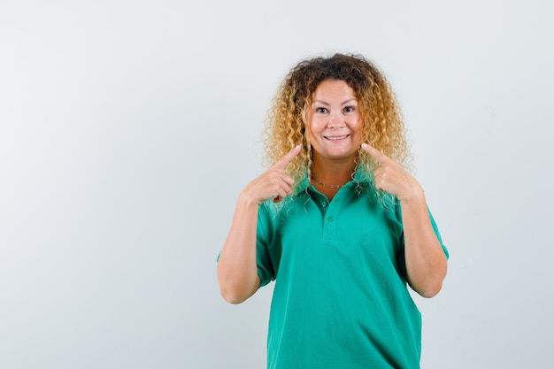 Portrait de jolie dame blonde pointant sur son sourire en t-shirt de polo vert et à la vue de face joviale