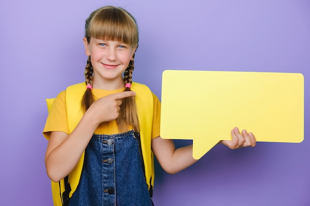 Portrait d'une jolie écolière caucasienne positive pointant sur une bulle jaune, porte un sac à dos, posant isolée sur fond violet en studio avec espace de copie pour le contenu promotionnel. Maquette