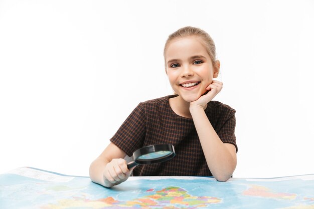 Portrait d'une jolie écolière regardant la carte du monde à travers une loupe tout en étudiant la géographie à l'école isolée sur un mur blanc