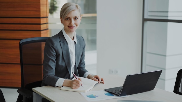 Portrait d'une jolie femme d'affaires blonde assise à une table écrivant dans un cahier souriant à la caméra