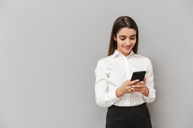 Portrait de jolie femme d'affaires avec de longs cheveux bruns en tenue de soirée souriant et à l'aide de téléphone mobile, isolé sur mur gris