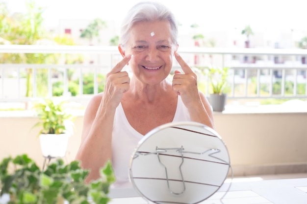 Portrait d'une jolie femme âgée aux cheveux gris appliquant une crème anti-âge sur son visage ridé