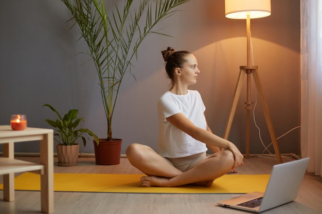 Portrait d'une jolie femme athlétique portant un t-shirt blanc pratiquant le yoga et l'étirement du corps à l'aide d'un ordinateur portable pour un cours en ligne assis dans une pose de lotus se détournant en regardant vers l'avant