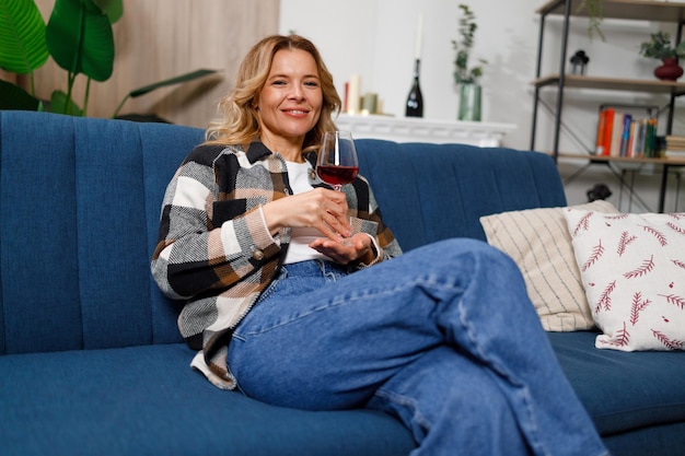 Portrait d'une jolie femme blonde d'âge moyen buvant du vin assise sur un canapé