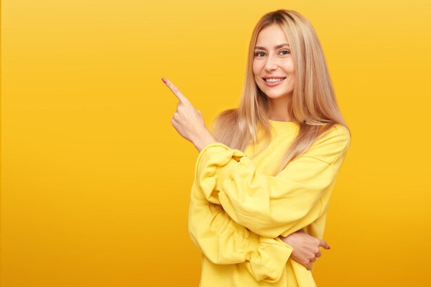 Portrait d'une jolie femme blonde souriante et pointant le doigt sur un espace vide sur fond jaune isolé