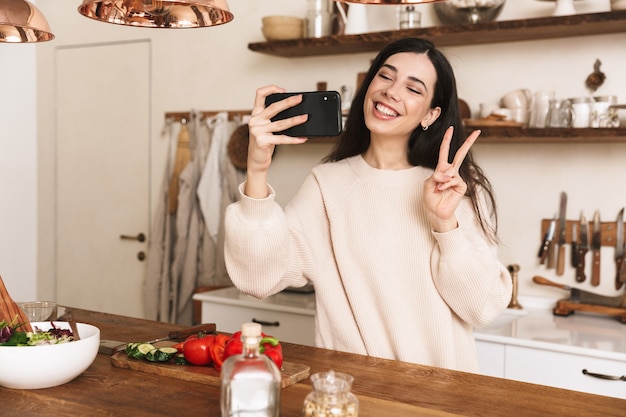 Portrait de jolie femme brune prenant selfie photo sur smartphone pendant la cuisson de la salade verte avec des légumes dans une cuisine élégante à la maison