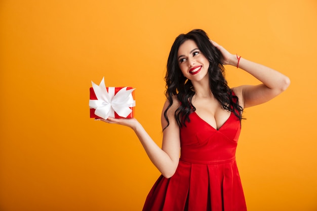 Portrait de jolie femme célibataire des années 20 en robe rouge profitant de sa boîte actuelle avec emballage cadeau, isolé sur mur jaune