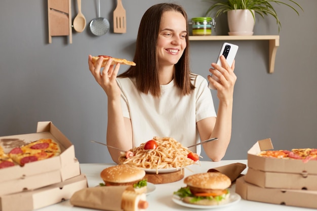 Portrait d'une jolie femme charmante aux cheveux bruns assise dans la cuisine et utilisant un téléphone portable tout en mangeant une pizza savoureuse en discutant avec des amis a laissé un service de livraison de commentaires positifs