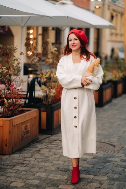 Portrait d'une jolie femme dans un béret rouge et un cardigan blanc avec des baguettes dans ses mains se promenant dans la ville d'automne