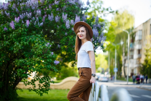 Portrait d'une jolie femme élégante dans un chapeau aux longs cheveux blonds dans le parc, vêtue d'un t-shirt blanc et d'un pantalon marron.