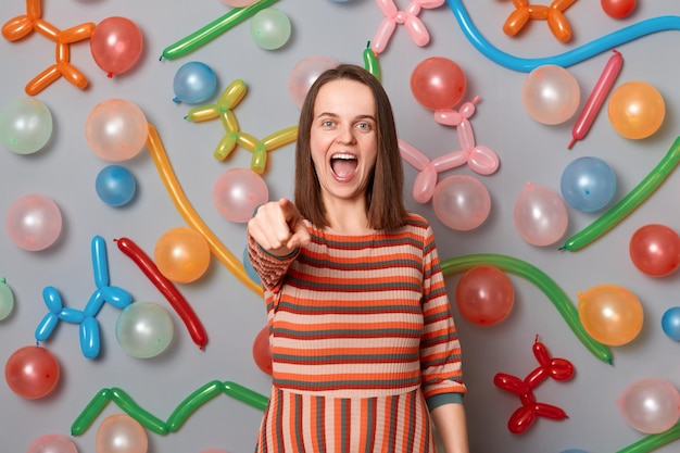 Portrait d'une jolie femme excitée aux cheveux bruns portant une robe rayée posant sur fond gris avec des ballons gonflés pointant vers la caméra vous invitant à sa fête d'anniversaire