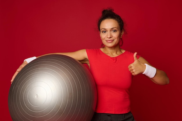 Portrait d'une jolie femme hispanique en forme tenant un ballon en forme et montrant un pouce vers le haut en regardant la caméra Copier l'espace libre pour le texte sur fond de couleur rouge