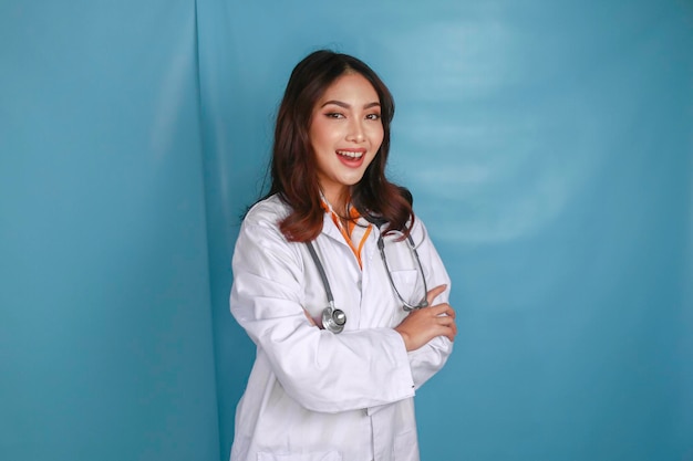 Portrait d'une jolie femme médecin souriante amicale et confiante, les bras croisés, porter un stéthoscope de laboratoire blanc isolé sur fond de couleur bleue