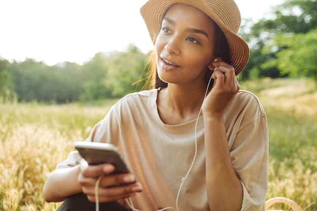 Portrait d'une jolie femme portant un piercing à la lèvre et un chapeau de paille écoutant de la musique sur un smartphone assis sur l'herbe dans un parc verdoyant