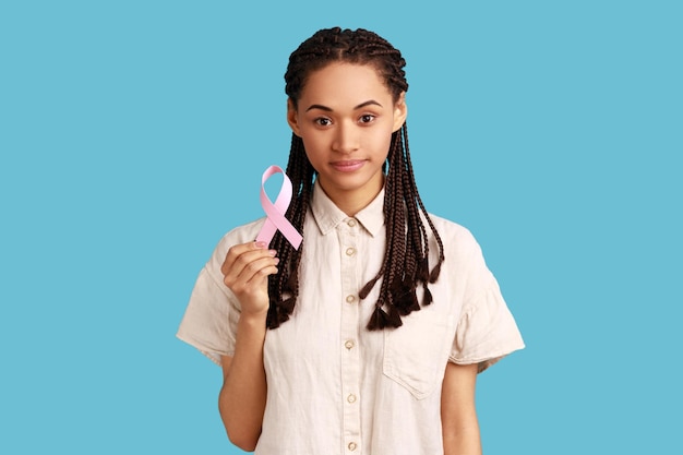 Portrait d'une jolie femme positive avec des dreadlocks noirs tenant un ruban rose, symbole de la sensibilisation au cancer du sein, portant une chemise blanche. Studio intérieur tourné isolé sur fond bleu.