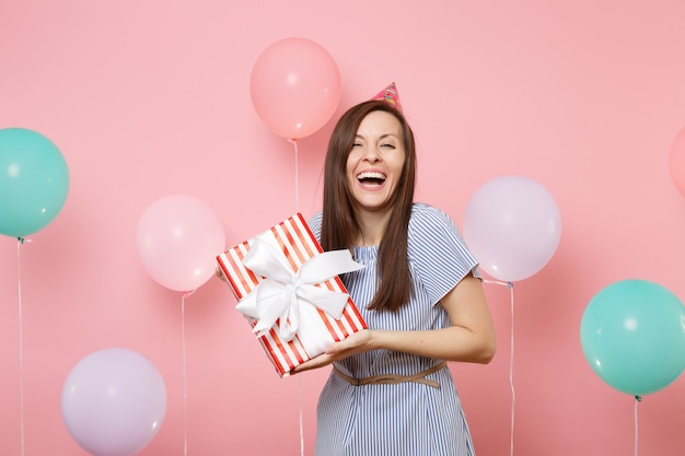 Portrait d'une jolie femme riante en chapeau d'anniversaire et robe bleue tenant une boîte rouge avec cadeau présent sur fond rose pastel avec des ballons à air colorés. Fête d'anniversaire, émotions sincères.