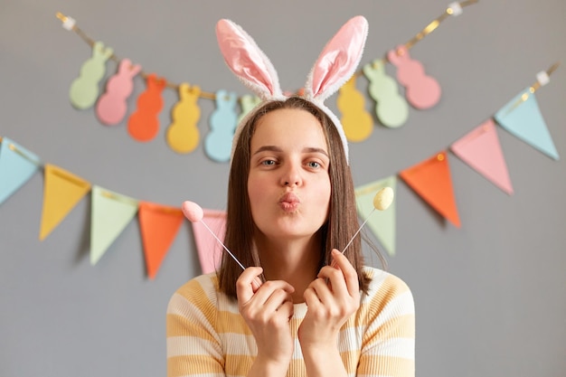 Portrait d'une jolie femme romantique portant des oreilles de lapin tenant un gâteau de Pâques coloré isolé sur fond gris décoré envoyant un baiser d'air félicitant pour les vacances