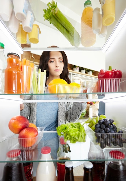 Portrait d'une jolie femme sérieuse debout près d'un réfrigérateur ouvert plein de légumes et de fruits sains