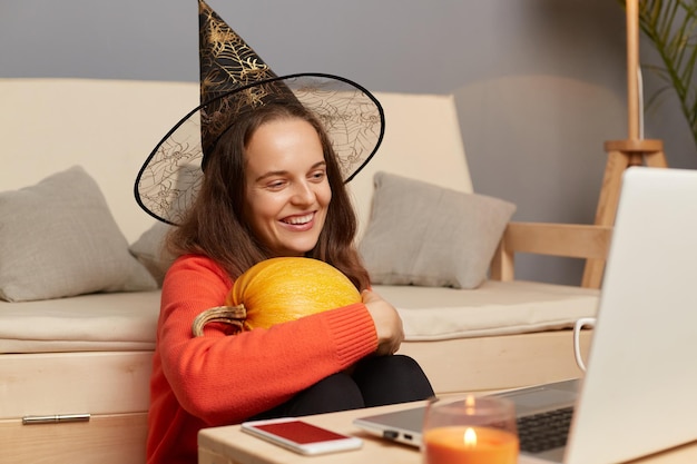Portrait d'une jolie femme souriante et heureuse portant un chapeau de sorcière devant un ordinateur portable à l'intérieur de la maison regardant l'écran de l'ordinateur portable avec le sourire embrassant la citrouille célébrant Halloween